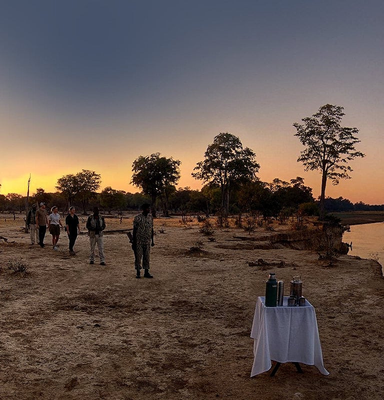 Walking safari group approaching a table with thermoses by a river in Zambia at sunset.
