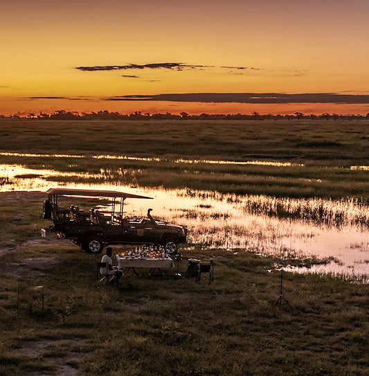 An open safari vehicle and a table setup next to a floodplain in the Chobe region at sunset.