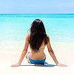 A woman sitting on white sand looking toward clear ocean waters.