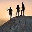 Silhouettes of a guide and two travelers standing on a rocky ridge at sunset.