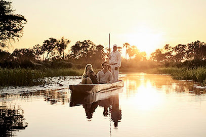 Travelers being poled along a calm wetland channel in a wooden mokoro boat at sunset.