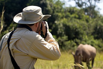 A photographer in a sun hat taking a picture of a rhino grazing in the grass.