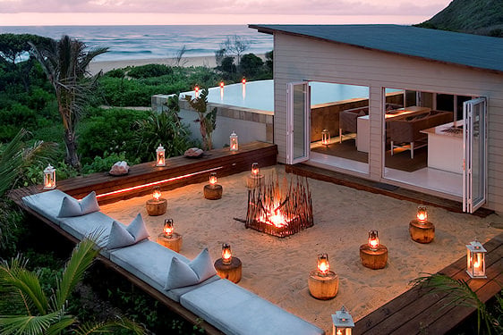 An illuminated open-air sand fire pit and lounge at a beach resort at dusk.