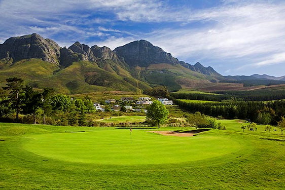 A golf green with a pin flag set against a backdrop of mountain slopes.