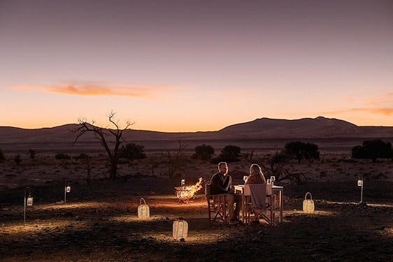 A couple seated for a private bush dinner near a fire pit in an arid landscape at sunset.