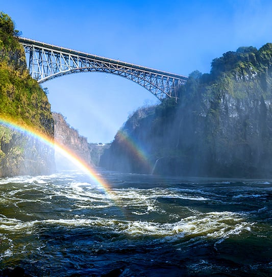 The Victoria Falls Bridge spanning the gorge above the Zambezi River with a double rainbow in the mist.