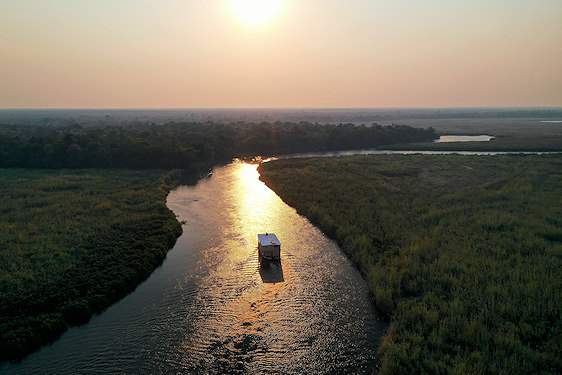 An aerial view of a safari boat moving down a river channel in the Okavango Delta at sunset.