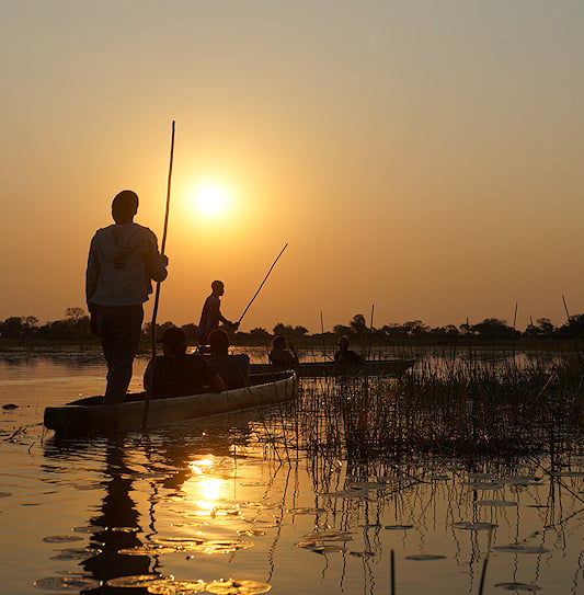 Tourists on a mokoro safari through the water channels of the Okavango Delta in Southern Africa at sunset.