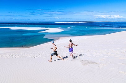 Two people running down a white sand dune toward shallow coastal lagoons in Mozambique.