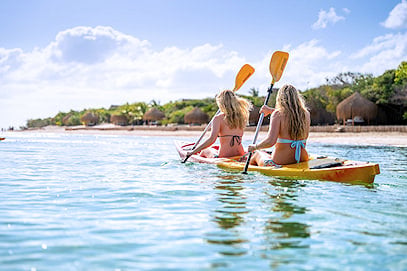 Two people paddling a yellow kayak in calm sea water near thatched beach huts.