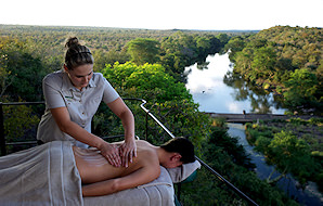 A woman enjoying a massage on an elevated outdoor patio with a captivating panorama of a river meandering through the African wilderness.