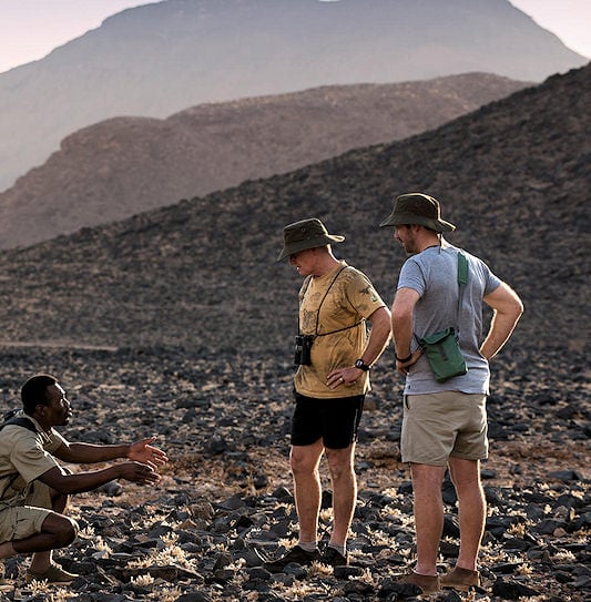 A safari guide crouching on a rocky desert landscape near Sossusvlei while talking to two tourists.