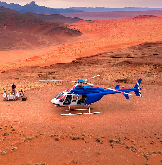A blue helicopter parked on a desert clearing next to a small outdoor dining setup by the red sand dunes of Sossusvlei.