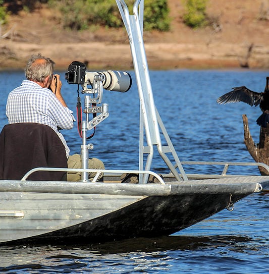 A man on a boat using a large telephoto lens to photograph a waterbird on the Chobe River.