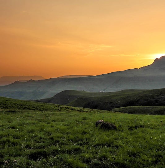 Green rolling hills and flat-topped peaks in the Drakensberg mountains under an orange sunset.