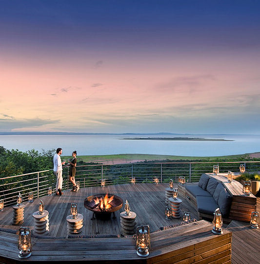 A man and a woman standing on the wooden viewing deck at Bumi Hills Safari Lodge, overlooking the lake at sunset.