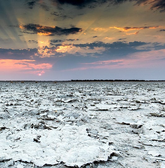Sunbeams radiating through dark clouds over the vast, cracked salt flats of Nxai Pan at sunset.