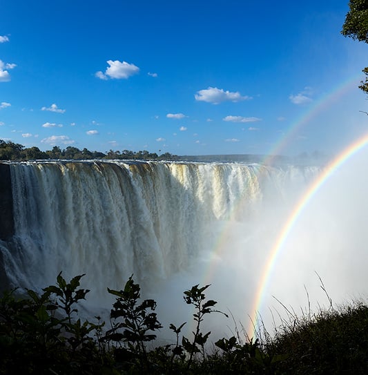 Victoria Falls in full flow, with a rainbow forming in the mist under a blue sky, Zimbabwe.