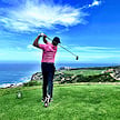 A golfer finishing a drive on an elevated tee box with panoramic ocean views.
