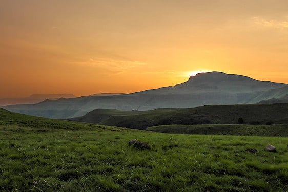 The sun setting behind a mountain ridge above green grasslands in the Drakensberg.