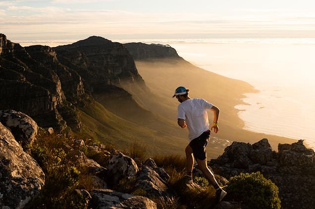 A runner on a rocky trail along Table Mountain overlooking the Cape Town coastline at sunset.