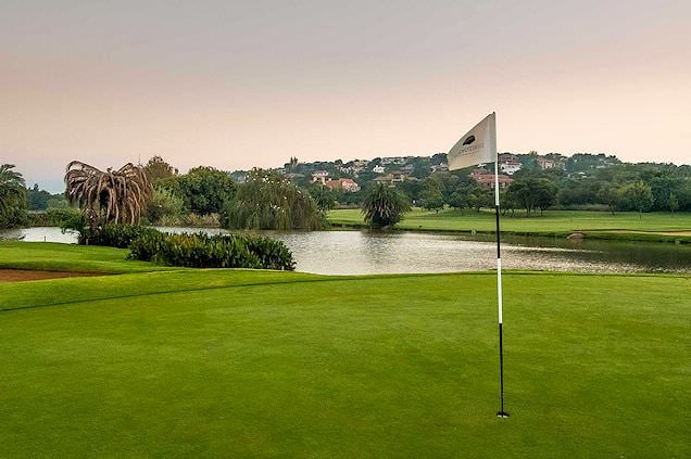 A golf green and flagstick beside a water hazard with houses on a hill in the distance.
