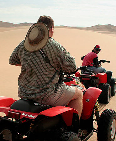 Two riders navigating red quad bikes across open desert sand dunes in Namibia.