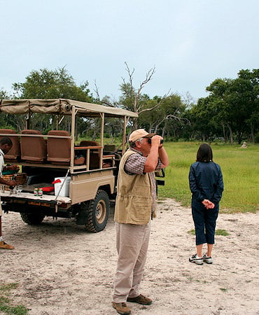 Safari-goers standing beside an open game drive vehicle while a man scans the landscape through binoculars.
