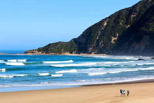 Hikers walking along a wide sandy shoreline below green coastal hills, Sedgefield, Garden Route, South Africa.