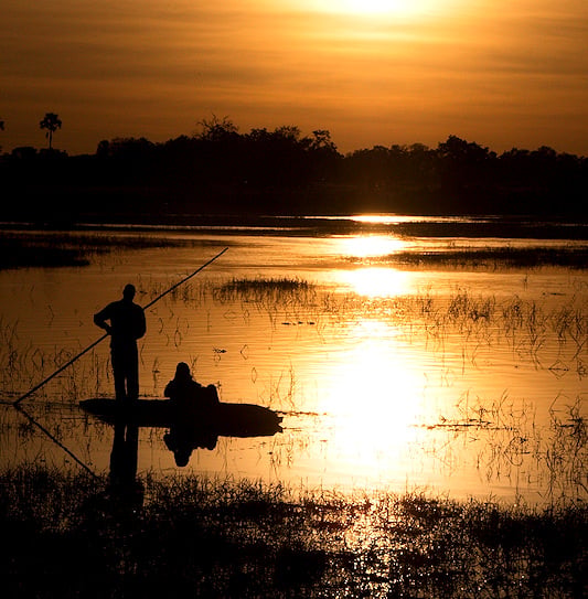 A silhouette of a person in a mokoro on the waterways of the Okavango Delta at sunset.