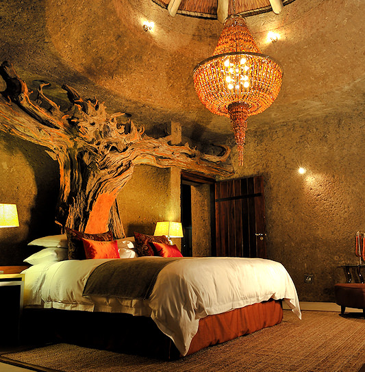 A bedroom suite at Sabi Sabi Earth Lodge featuring rough-textured walls, a tree-root headboard, and a large amber chandelier.