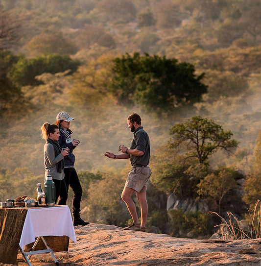 A safari guide and two guests standing on a rocky outcrop next to a picnic table, overlooking the bushveld in Kruger National Park.