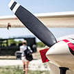 A close-up of a light aircraft propeller with a passenger walking onto a dirt airstrip.