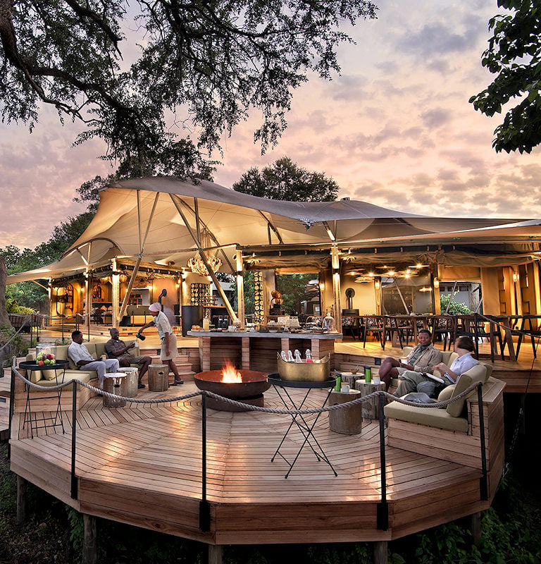 Guests relaxing in an outdoor lounge area with a central fire pit under a stretch canvas tent at dusk.