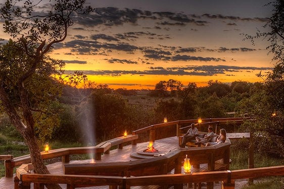 Guests gathering around a sunken fire pit on a timber veranda at dusk in the Okavango Delta.