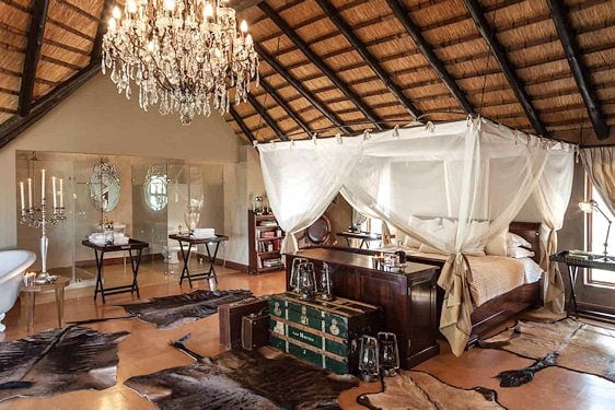 A thatched safari suite interior featuring a four-poster canopy bed, chandelier, and standalone bathtub.