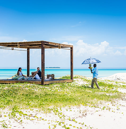 Staff member serving fresh fruit to seated guests under a timber shade canopy on Medjumbe Island.