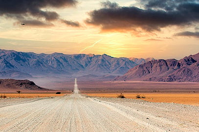 Long straight gravel road stretching across open desert toward rugged mountains, in Namibia.