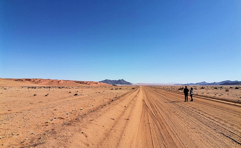 Two people standing on a gravel road in a desert landscape with red sand dunes, D707 road near Aus, Namibia