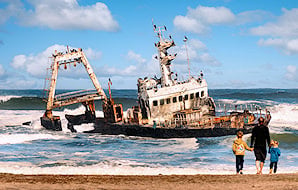 Family walking on the beach looking at a rusted shipwreck in the crashing waves along the Skeleton Coast.