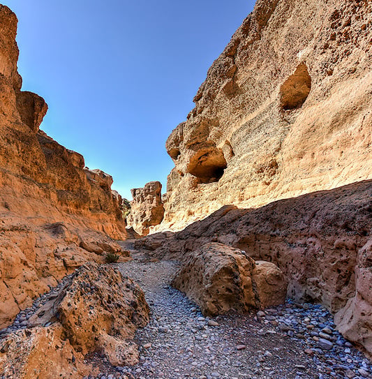 A stony path winding between the towering, eroded rock walls of Sesriem Canyon under a clear blue sky.