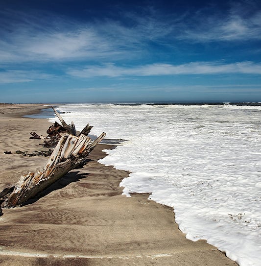 Ocean waves and white foam washing onto a sandy beach next to a piece of weathered wreckage on the Skeleton Coast.