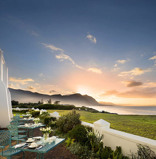 Outdoor patio breakfast tables at The Marine Hotel in Hermanus, overlooking Walker Bay and coastal mountains at sunrise.