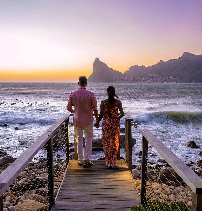 Couple holding hands on a wooden boardwalk at sunset, looking out at the ocean with Chapman's Peak in the background in Cape Town.