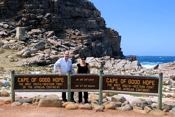 A couple posing behind the wooden Cape of Good Hope sign on the rocky coastline.