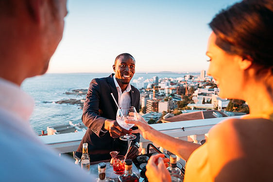 Guests receiving drinks from a bartender at an outdoor rooftop bar overlooking the ocean at dusk.