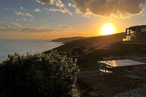 An outdoor dining terrace on a grassy hill overlooking the ocean coastline at sunset.