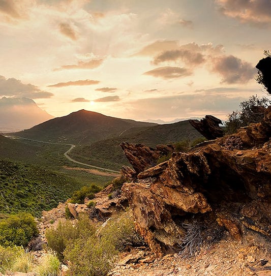 A road through a valley during sunset in the Klein Karoo, with rock formations in the foreground.