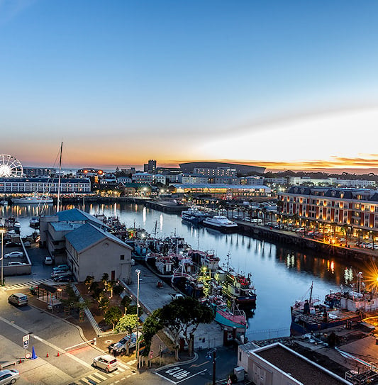 Boats docked in a harbor canal lined with illuminated buildings and a Ferris wheel at dusk, V&A Waterfront, Cape Town.