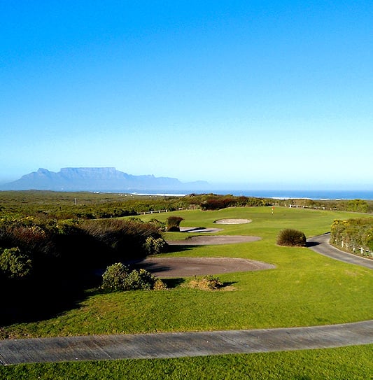 A cart path alongside sand traps and vegetation with Table Mountain across the bay in Cape Town.